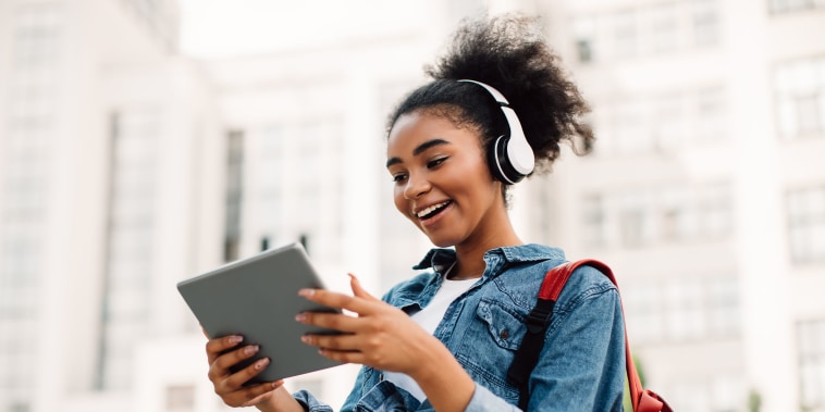 Excited African American Student Girl Using Digital Tablet Wearing Headphones Standing Near University Building Outdoors