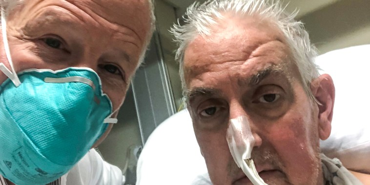 Two white men with white hair take a selfie photo. one is in a white lab coat and facemask while the other sits in a hospital bed with a tube connected to his nose.