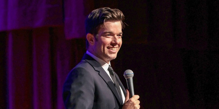 John Mulaney smiles in a suit while holding a microphone on stage in front of a red/purple curtian.