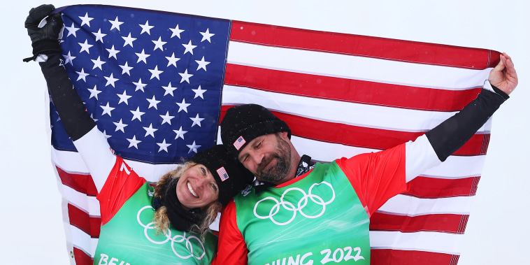 Snowboard - Beijing 2022 Winter Olympics Day 8 - Two people stand in front of snow holding an American flag and smiling. They are in snow gear and propped their snowboards in front of them.