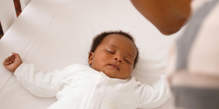 A Black baby dressed in a white onesie sleeps in a crib as a parent looks over.