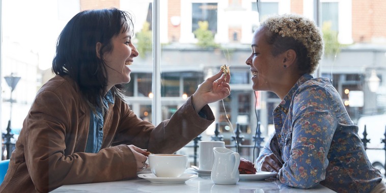 Happy young woman feeding food to girlfriend sitting in cafe