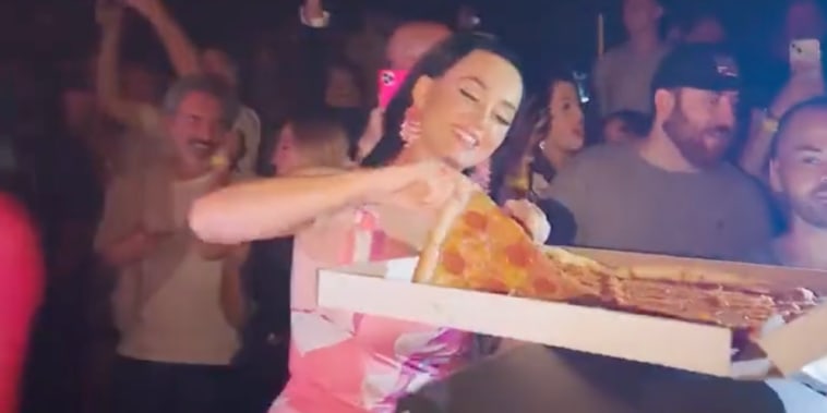 Katy Perry smiles as she pulls a piece of pizza from a box in front of a crowd