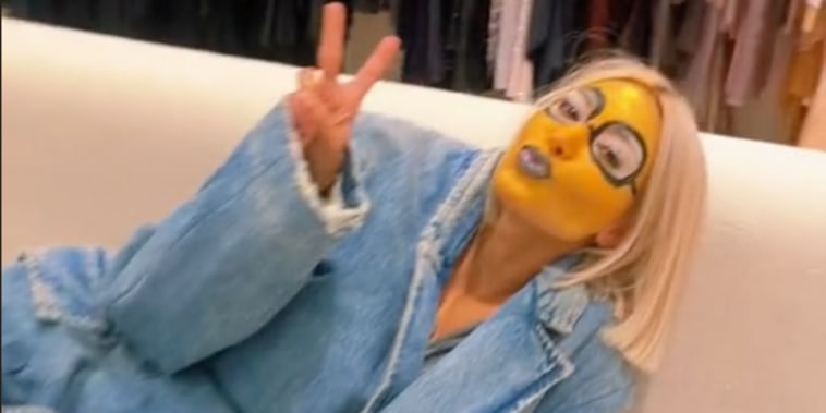 Kardashian poses in a jean blazer on a white couch with yellow face paint and black painted on glasses. her hair is styled in a blond bob.