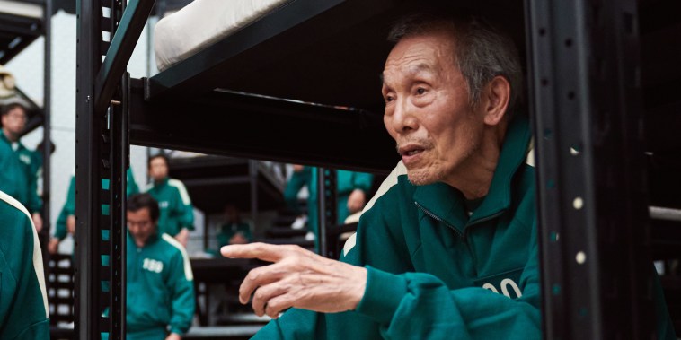An older man in a numbered green tracksuit sits on a bunk bed, looking off-camera and pointing straight ahead. 