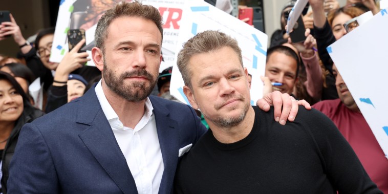 Ben Affleck (left) and Matt Damon stand in front of a cheering crowd with "Air" posters. Affleck is in a suit and collared shirt, Damon is in a black long sleeved crew neck sweater.
