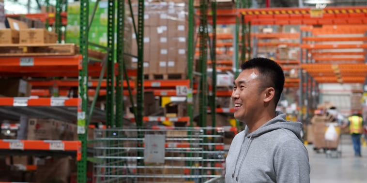 A young Asian man stands in front of shelves in a warehouse.