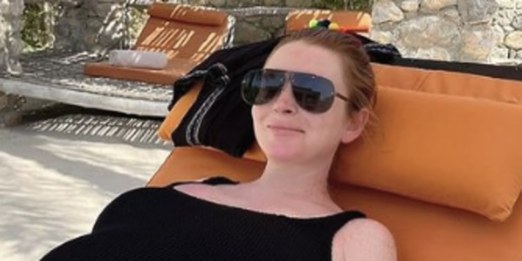 Lohan wears black aviator sunglasses and a black one-piece bathing suit to recline on an orange lounge chair.