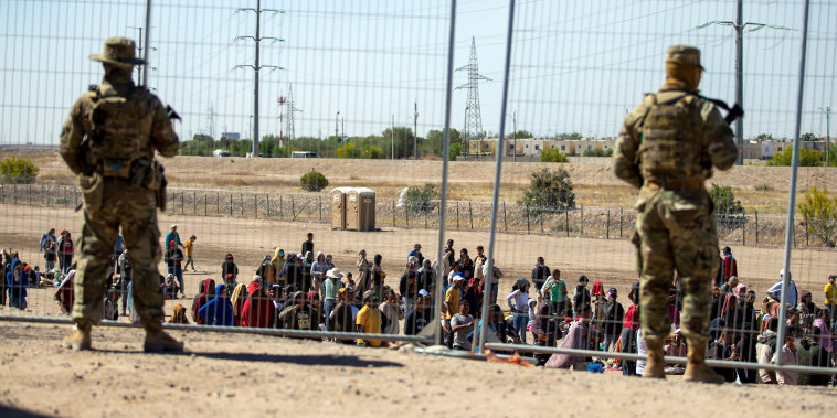 Migrantes esperan en fila junto a la valla fronteriza bajo la vigilancia de la Guardia Nacional de Texas para entrar en El Paso, el miércoles 10 de mayo de 2023.