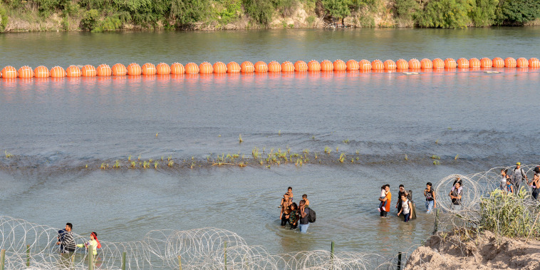 Migrants walk by a string of buoys placed on the water along the Rio Grande border with Mexico in Eagle Pass, Texas, on July 16, 2023. The buoy installation is part of an operation Texas is pursuing to secure its borders, but activists and some legislators say Governor Greg Abbott is exceeding his authority.
