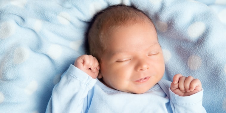 Newborn Baby Sleeping Smiling. Cute Infant Child in Wrap Bodysuit. New Born Little Boy smile in Blue Clothes lying on Blanket. Small Kid Face Close up Portrait holding Hands First