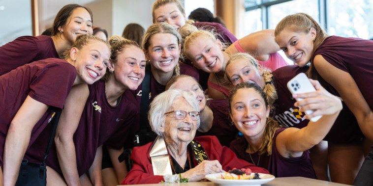 Sister Jean sits at a table surrounded by young women in maroon loyola shirts as they take a selfie.