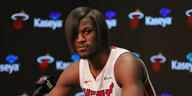 Butler in a Miami Heat jersey speaks into a microphone. His hair has been straightened and his bangs cover his right eye. He also has several facial piercings.