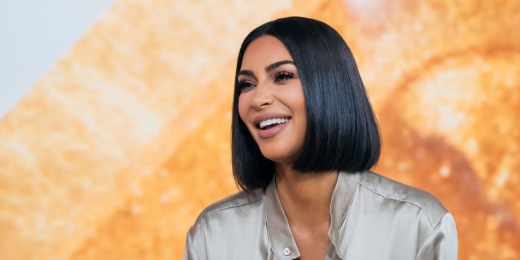 kim kardashian laughs and smiles at something off camera in a taupe silk shirt in front of a sparkly gold background. her hair is cut into a short brown bob.