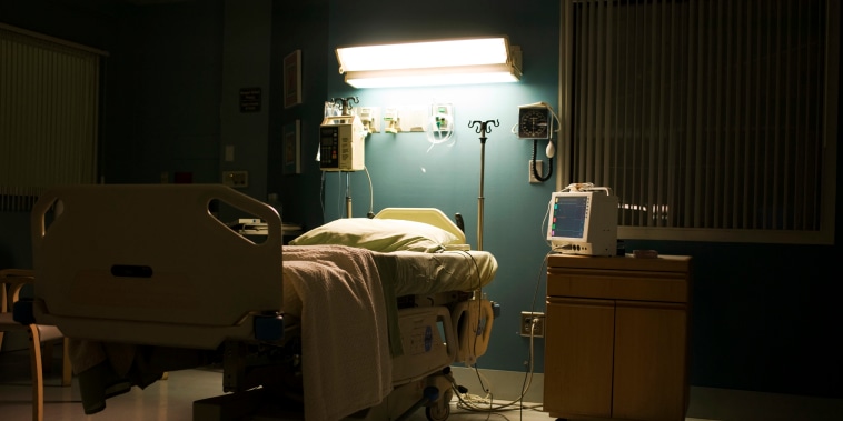 A bed sits under a single light in a dark hospital room.