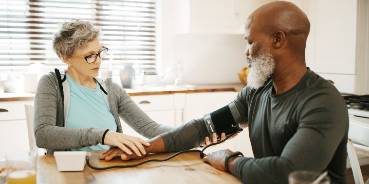 Senior woman sitting and checking her husband's blood pressure in their kitchen