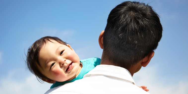 An East Asian baby boy smiling on his father's shoulder.