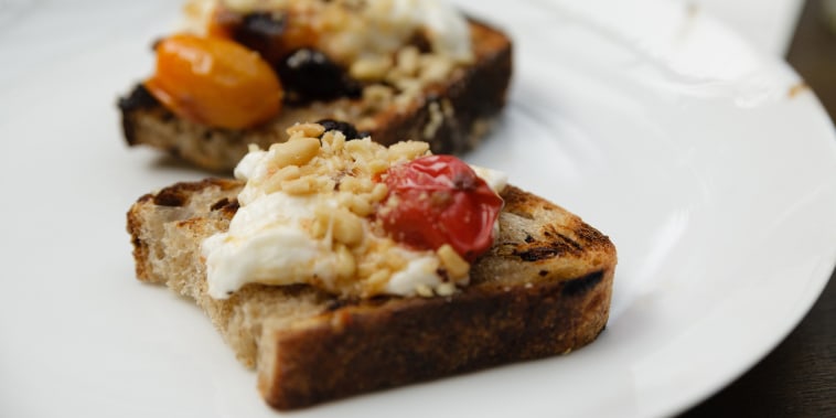 Burrata toasts with cherry tomatoes, olives and pinenuts on a white plate.