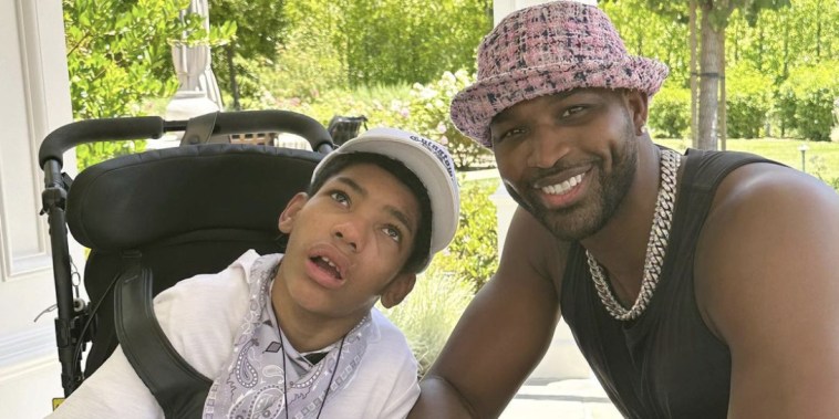 Amari sits in a wheelchair wearing a white T-shirt and white baseball cap. On the right, Tristan Thompson smiles at the camera wearing a pink tweed bucket hat and black tank top.