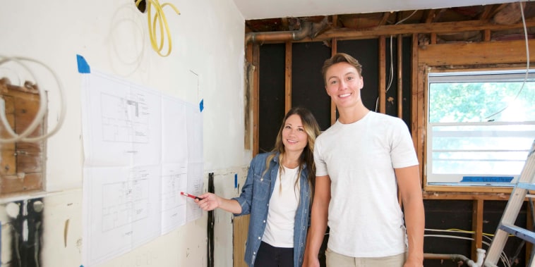 Two women smile at a camera inside a home under construction. On their right, one points at an architectural rendering.