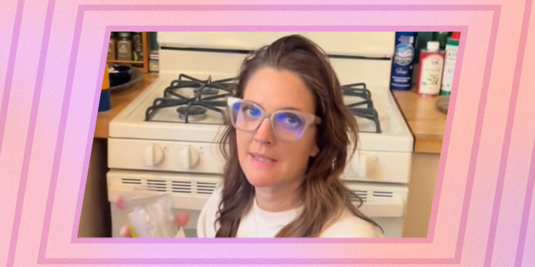 Drew Barrymore in glasses kneels on her kitchen floor in front of a white gas stove.