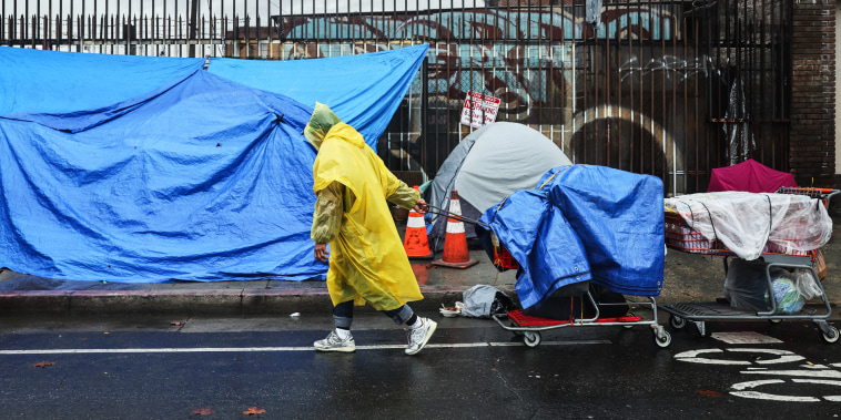 A person walks with carts in the rain near an encampment of unhoused people