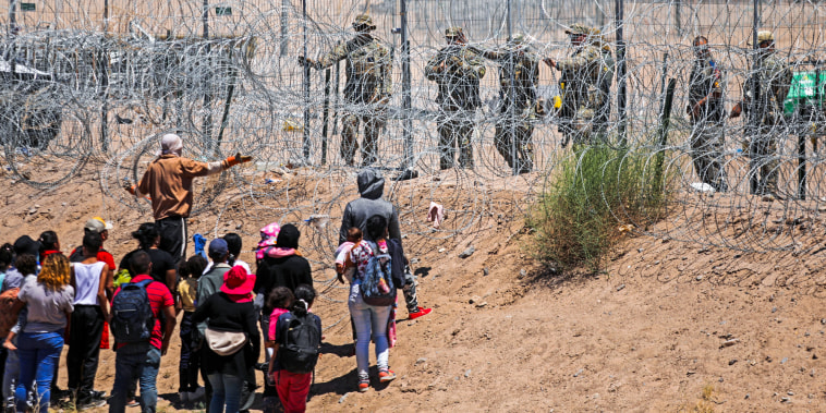 Migrants stand in front of a barbed wire fence opposite from National Guard agents