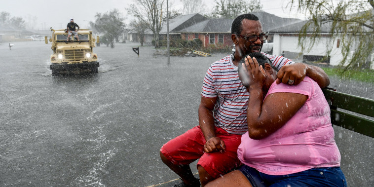 Two people embrace while sitting on a bench as a rain shower soaks them outside, as a volunteer evacuation truck is in the distance