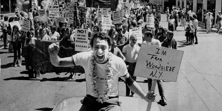 Harvey Milk sitting on top of a car during the Pride Parade. He is holding a placard that reads: 'I'm from Woodmere, NY'. 