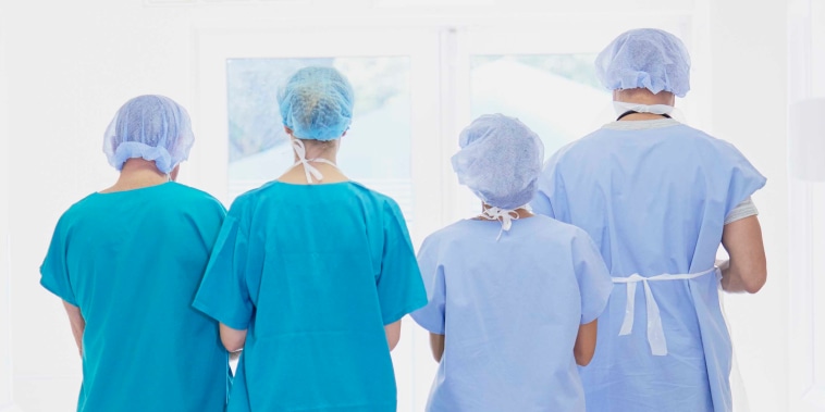 Rear view of four medical staff wearing scrubs walking in hospital corridor.