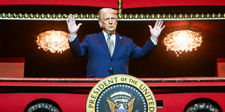 Donald Trump, with both hands raised, stands on a balcony inside of a building