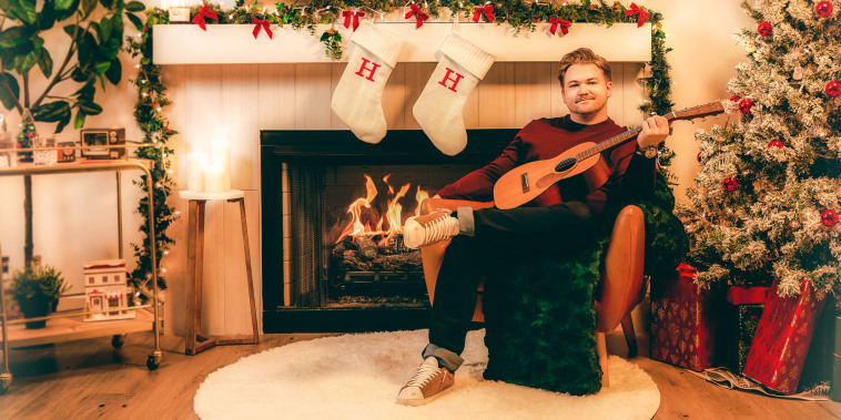 Hunter Hayes sits on chair in a living room holding a guitar, with the fireplace going and Christmas tree lights on