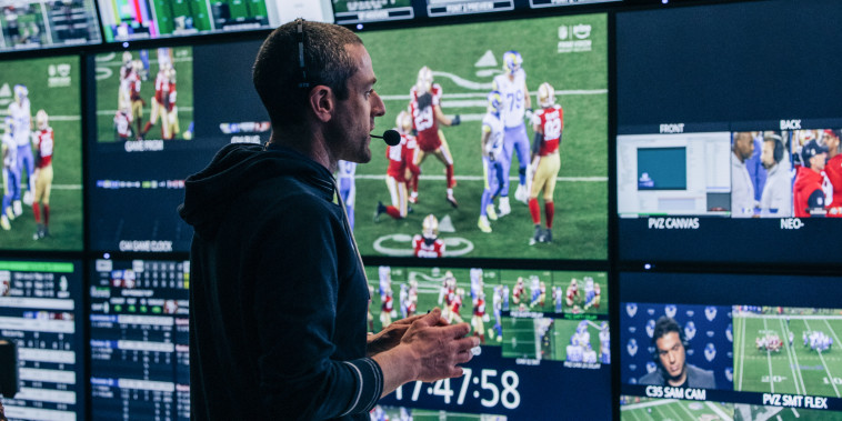 A person inside of a control room looks at various television screens stacked atop one another displaying different angles of a football game