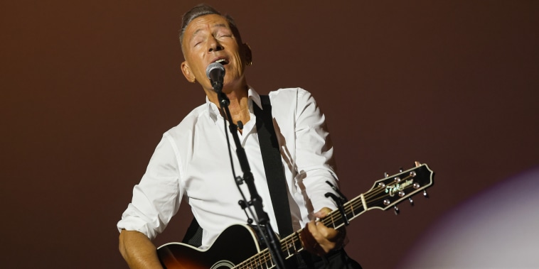 Bruce Springsteen sings into a microphone while playing an acoustic guitar.