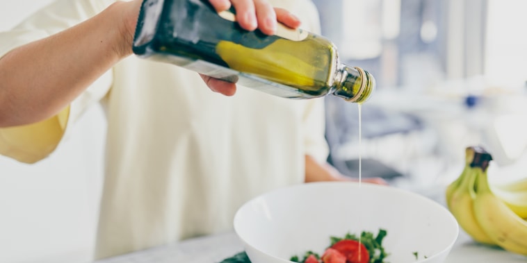 Hands pouring olive oil into a white bowl with chopped tomatoes and leafy greens. Fresh vegetables around.
