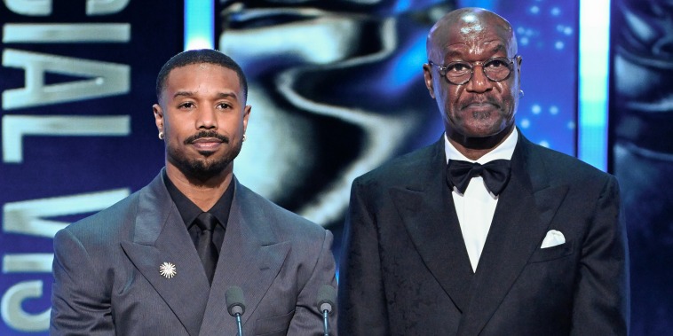 Michael B. Jordan and Delroy Lindo stand at a lectern on stage during an award ceremony. They look nonplussed.