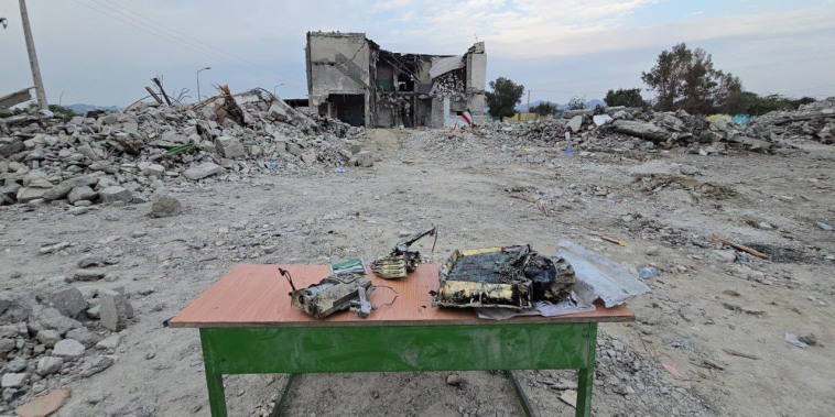 Missile debris is gathered on a table outside, a destroyed building is seen is the distance