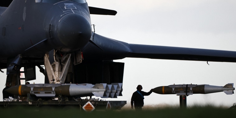 A soldier wearing earmuffs with one hand on a large missile is silhouetted against the sky.