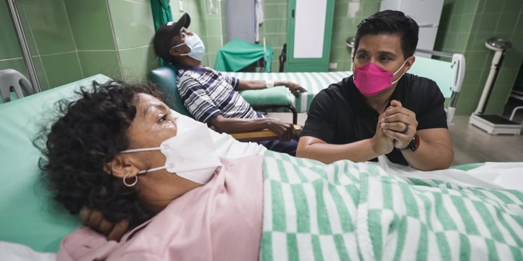George Solis, right, wearing a pink mask, speaks with Yonelkys García who is laying in a hospital bed and wearing a white mask