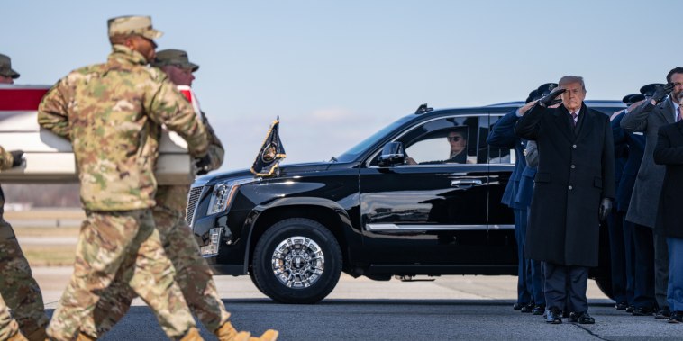 Army members carry a casket across an aircraft tarmac as Donald Trump and company stand to watch while saluting