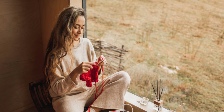 Young Woman Holding Needles and Knitting Handmade Piece From Red Yarn while Sitting near Window.