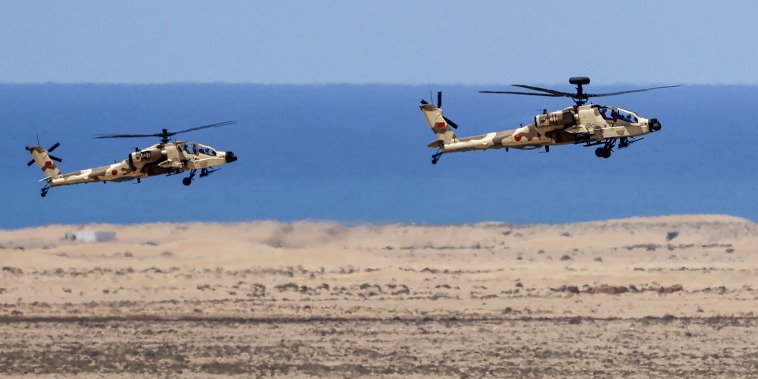 Two camouflage helicopters hover over a desert landscape with blue waters in the background.