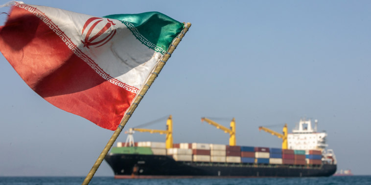 An Iranian flag appears in the foreground with a cargo ship in the background.