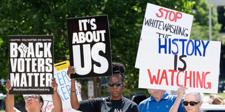 People hold signs that say "Black Voter Matter", "It's About Us", and "History Is Watching" while standing outside