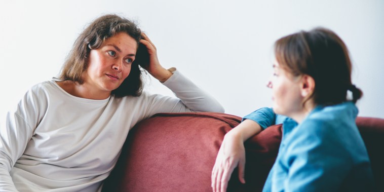 Two women in conversation on a red sofa in a modern living room. Woman in white shirt looking thoughtfully at the woman in a blue shirt. Indoor plant in the background.