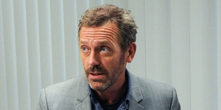 Hugh Laurie sits inside of a doctor's office next to medical supplies as he speaks to a patient sitting in front of him