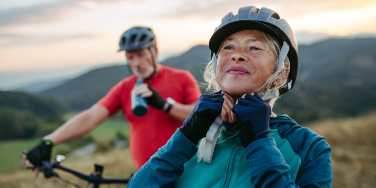 Portrait of beautiful senior couple on bike ride in autumn nature. Taking break, drinking water from sport bottle and fastening helmet.