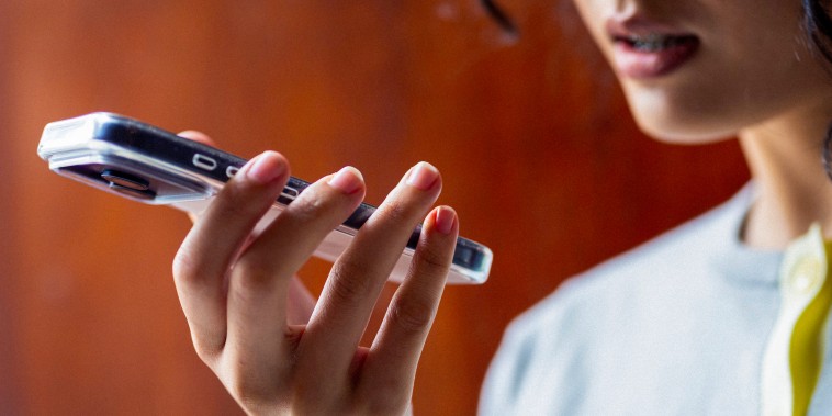 Young woman holds a smartphone to speak a voice message, focused expression and warm indoor lighting.