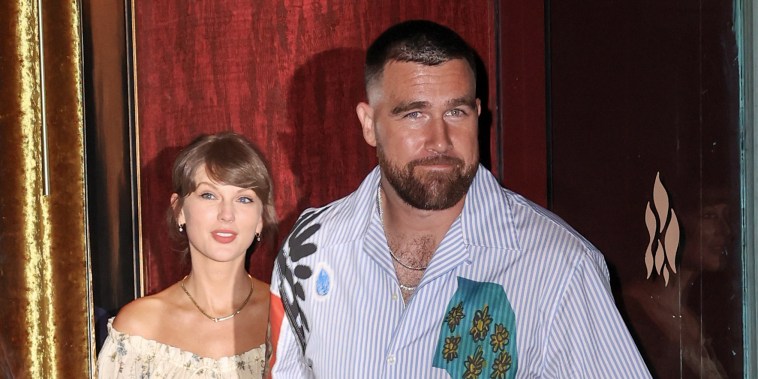 Travis Kelce, looking toward the camera, holds Taylor Swift's hand as they exit a building to the street.