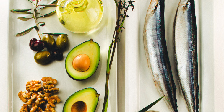Still life of foods like olives, canola oil, mackerels, flaxseeds, avocado, and walnuts. 